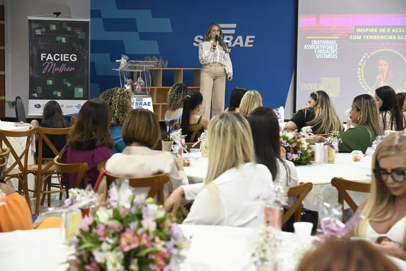 Rejane Duarte de pé, com o microfone, falando a mulheres sentadas em mesas com flores, diante de um painel do Sebrae