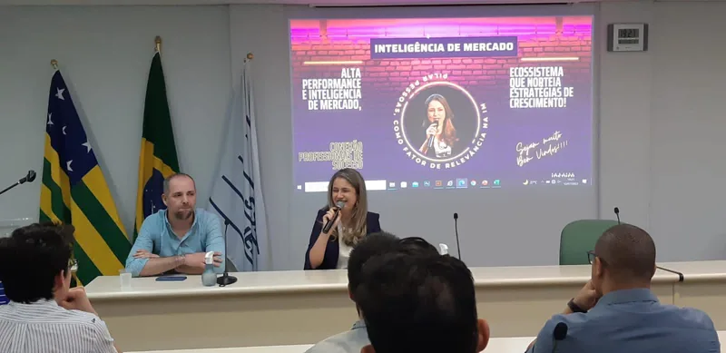 Rejane Duarte sentada à mesa de um painel, falando ao microfone ao lado de outro convidado, com um telão ao fundo