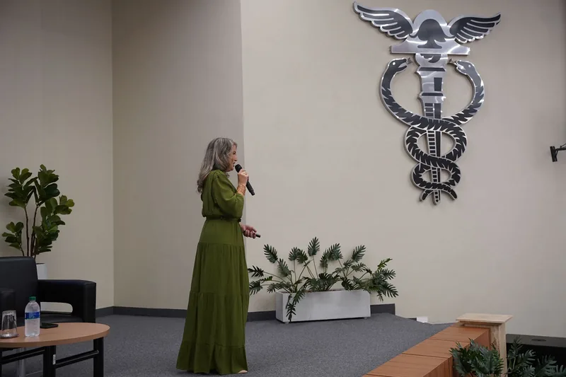 Rejane Duarte no palco de um auditório, de vestido verde, falando ao microfone