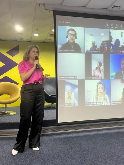 Rejane Duarte de pé, com o microfone, ao lado de um telão com participantes numa videochamada